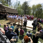 Ludzie zgromadzeni wokół drewnianej sceny, na której tańczą ludzie w góralskich strojach, z tyłu drzewa.