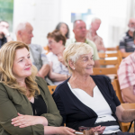 Na pierwszym planie siedzące na krzesłach dwie kobiety, z tył inni ludzie.