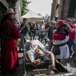 Leżący na machinie do tortur mężczyzna, wokoło niego zebrani ludzie.