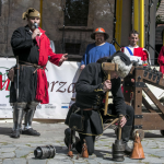 Mężczyźni w strojach średniowiecznych, mężczyzna uderzający młotkiem, z tyłu scena.