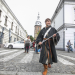 Mężczyzna w stroju z epoki średniowiecza idący brukowaną ulica, za nim inne osoby, po bokach budynki, z tyłu kościół.