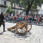 Mężczyźni w zbrojach rycerskich odpalają działo, przez nimi zgromadzona na placu publiczność.