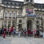 Grupa ludzi w historycznych strojach zgromadzona na rynku na tle ratusza.