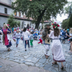 Tańczące na placu dziewczęta, z tyłu publiczność, drzewo, budynki.