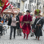 Grupa osób w historycznych strojach idąca deptakiem, obok kobieta w stroju współczesnym.