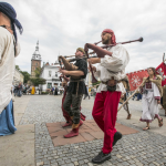 Grupa osób w historycznych strojach idąca ulicą, na pierwszym planie mężczyźni grający na dawnych instrumentach.