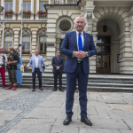 Mężczyzna w garniturze, za nim dwóch mężczyzn w garniturach, po bokach osoby w strojach z epoki średniowiecza, wszyscy na tle ratusza.