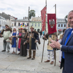 Klaszczący mężczyzna w garniturze, za nim grupa ludzi w strojach historycznych, jeden trzyma sztandary, wszyscy zgromadzeni na rynku.