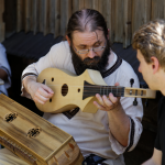 Grający na małej gitarze mężczyzna, obok dwóch innych mężczyzn.