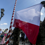 Żołnierz wciąga biało-czerwoną flagę na maszt, obok niego inny żołnierz, za nim ludzie.