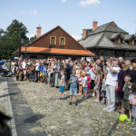 Dzieci i dorośli zgromadzeni na rynku miasteczka, w tle budynki.