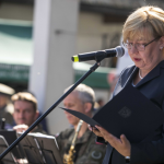 Mówiąca do mikrofonu kobieta czytająca tekst, za nią orkiestra.