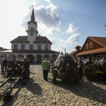 Sprzęt wojskowy i ludzie zgromadzeni na rynku miasteczka, po bokach budynki i budynek ratusza.