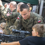 Mężczyźni w mundurach, przed nimi karabiny i oglądający karabin chłopiec.