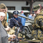 Żołnierze pokazują sprzęt wojskowy starszemu panu, za nimi mężczyzna w stroju przedwojennego policjanta.