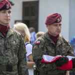 Mężczyźni w wojskowych mundurach, jeden z nich trzyma złożoną biało-czerwoną flagę.