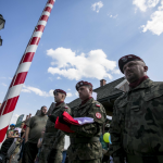 Mężczyźni w wojskowych mundurach, mężczyzna w środku trzyma złożoną biało-czerwoną flagę, przed nimi biało-czerwony maszt, za nimi dorośli i dzieci.