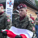 Mężczyźni w wojskowych mundurach, jeden z nich trzyma złożoną biało-czerwoną flagę.