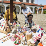 Siedząca kobieta i dwóch stojących mężczyzn za stołem z rękodziełem.