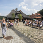 Idąca przez rynek miasteczka kobieta, za nią ludzie siedzący na ławkach.