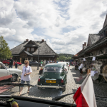 Zabytkowe auta widziane przez szybę samochodu, zatknięta w środku biało-czerwona flaga, w około budynki.