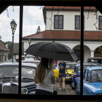 Kobieta pod parasolem, obok samochody i budynki, widziane przez okno budynku.