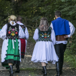 Idące polną drogą trzy kobiety w strojach regionalnych, z nimi mężczyzna, wszyscy odwróceni tyłem.