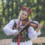 Grająca na skrzypcach dziewczyna w białej koszuli i ozdobnym gorsecie, w wianku na głowie.