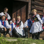 Grupa osób w strojach regionalnych u progu drewnianego domu.