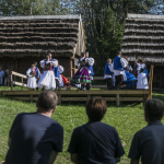 Tańcząca grupa osób w strojach regionalnych, przed nimi troje ludzi siedzących na ławce.