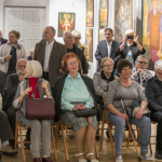 Grupa ludzi w sali; część siedzi na krzesłach, pozostali stoją za nimi.