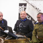 Trzech mężczyzn siedzących przy stoliku w mundurach policjantów, za nimi baner.