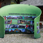 Duży zielony baner z napisem Nowatorska Małopolska Wieś 2019, pod spodem zbiór zdjęć.