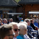 Publiczność siedząca na krzesłach na rynku miasteczka.