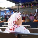 Dwie kobiety w białych czepcach z czerwonym haftem, w tle zespół regionalny na scenie.