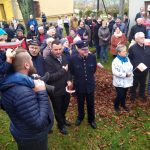 Grupa osób z biało czerwonymi chorągiewkami na tle budynków.