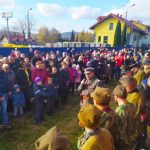 Mężczyzna w mundurze z mikrofonem przed grupą osób. Za jego plecami kilkunastu mężczyzn w mundurach.