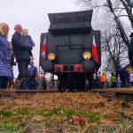 Lokomotywa na torach obok mężczyzna trzyma dziecko na rękach i kobieta. W tle grupa osób.