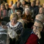 Grupa ludzi siedzących na krzesłach na pierwszym planie dwaj mężczyźni, jeden przegląda kalendarz, drugi z głową wspartą na dłoni.