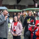 Mężczyzna przemawiający przez mikrofon, obok dziewczynka i kilka osób zebranych wokół.