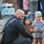 Kucający mężczyzna który trzyma w objęciach dziecko, obok stoi drugie.