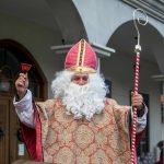 Mężczyzna przebrany w strój Świętego Mikołaja z rękami wzniesionymi ku górze. W jednej dłoni trzyma laskę w drugiej dzwonek.