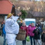 Kolędnik w białym stroju i słomianym kapeluszu trzymający drewnianą beczkę oraz gałązki jedliny.