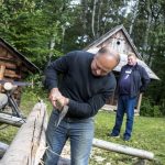 Mężczyzna tnący siekierą drzewo, z tyłu inny mężczyzna, po bokach drewniane domy.