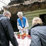 Chłopiec w szarej koszuli i niebieskiej kamizelce, w czapce na głowie, na tle starej chaty, przed stołem z przyprawami i ziołami. Przed nim kobieta i mężczyzna.