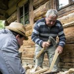 Starszy mężczyzna tnący siekierą drewnianą deseczkę. Obok starszy mężczyzna w kapeluszu.