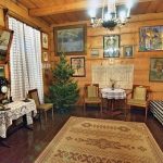 Wnętrze drewnianego domu, na ścianach wiszą obrazy, w kącie zielony świerk.