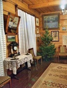Wnętrze drewnianego domu, na ścianach wiszą obrazy, w kącie zielony świerk.