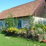 Drewniana chałupa od frontu otoczona kwiatami.