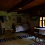 Fragment izby w drewnianej chałupie. Łóżko, stół, obrazy.